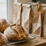 Bread rack holding sourdough boules and baguettes beside folded brown paper bags